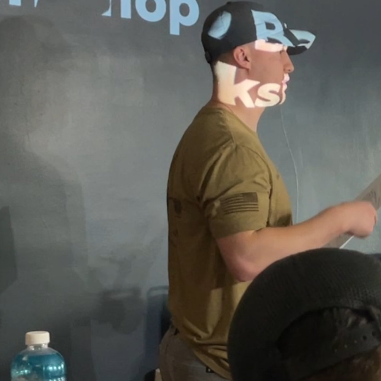Person wearing a cap with text, standing in front of a screen with projected text.